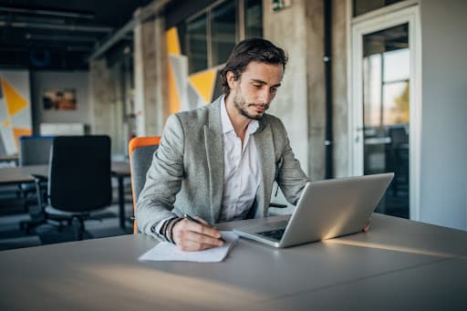 A young handsome businessman working on his laptop while jotting down notes at his office.