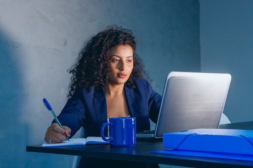 A woman wearing blue working on her laptop while jotting down notes. A blue cup and a blue envelope found on the table.